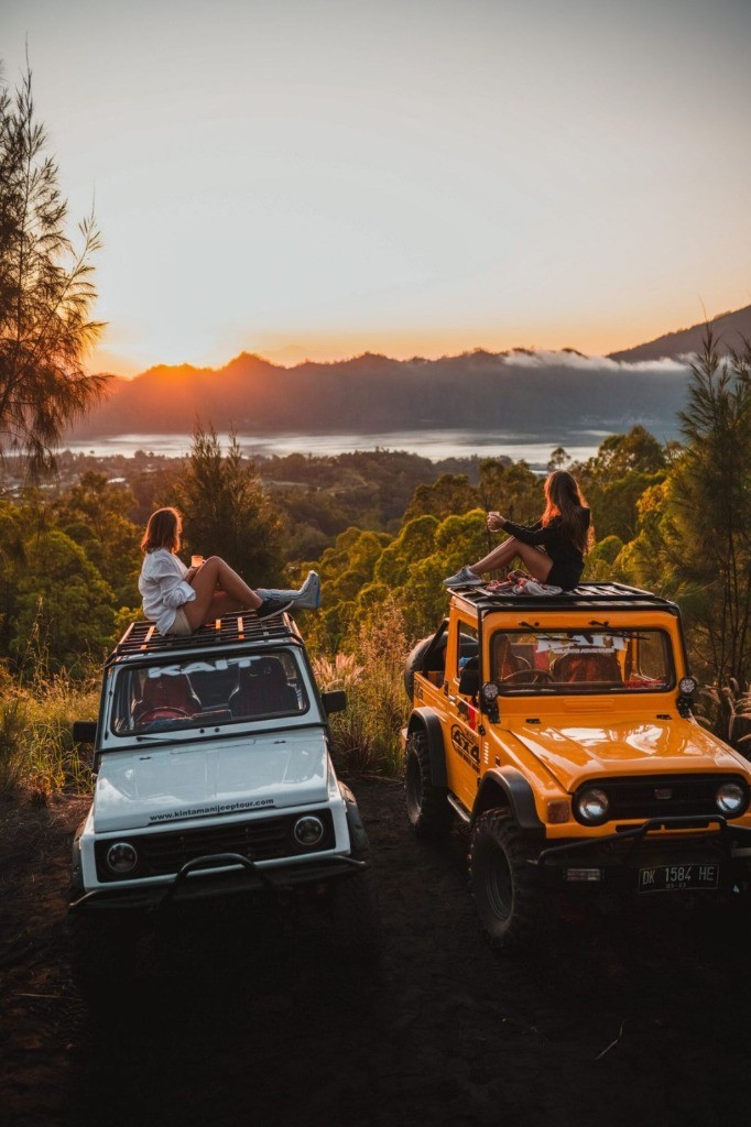 Two travelers sitting on classic 4x4 jeep rooftops overlooking Mount Batur caldera lake at sunrise